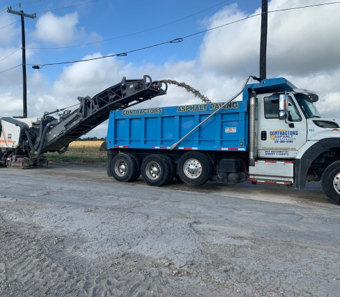On a cloudy day, a construction vehicle from Contractor's Asphalt is busy transferring asphalt from the road into a dump truck, ensuring smooth paving operations.