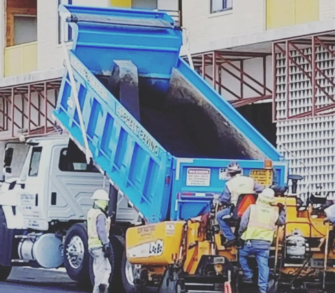 Workers in safety gear operate machinery near a blue dump truck unloading materials for an asphalt paving project on the construction site.