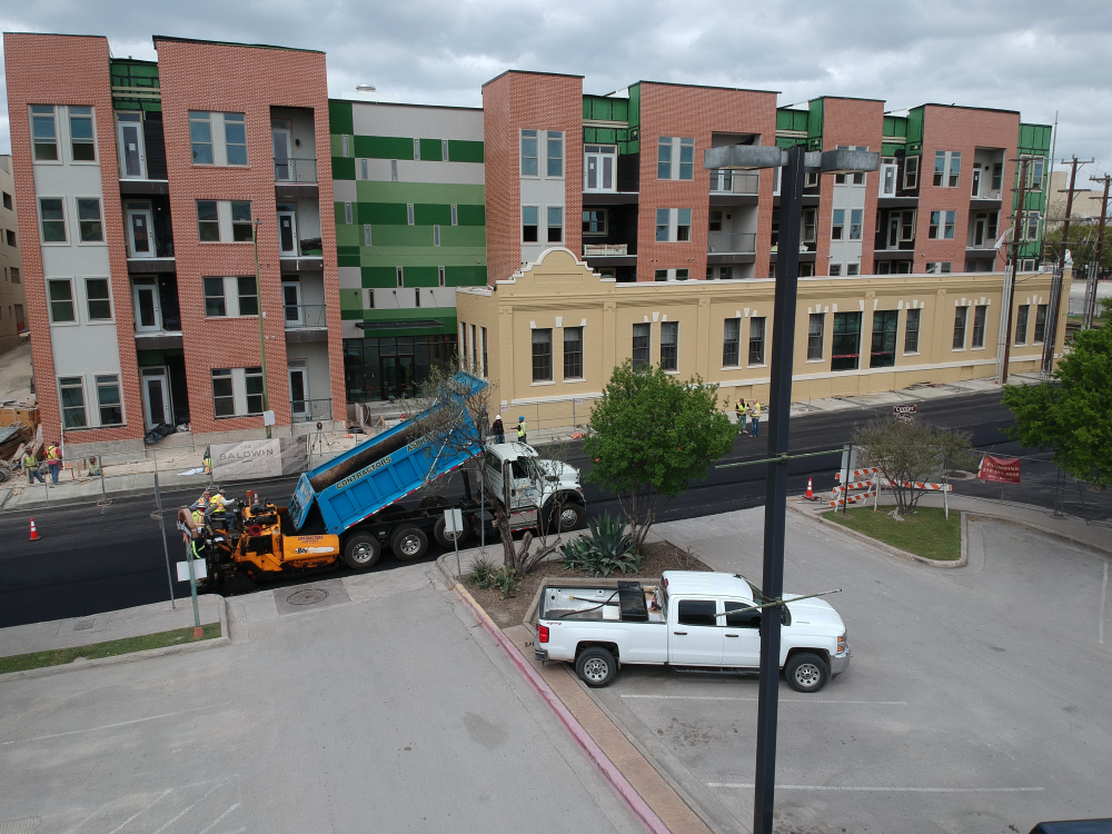 A paving crew from Contractor's Asphalt works diligently on a road in front of a multi-story residential building. A dump truck unloads asphalt into machinery for the asphalt paving project, while workers skillfully direct traffic. A white pickup truck is parked nearby.