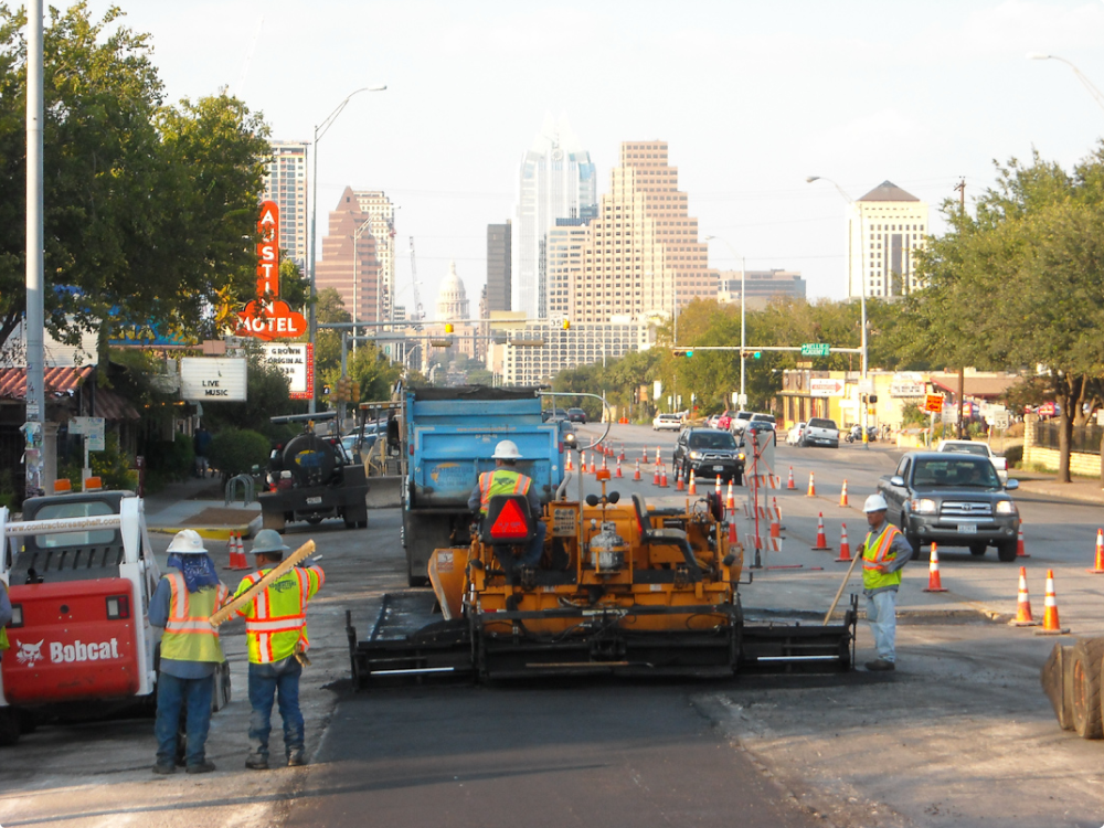A bustling road construction scene unfolds with workers, vehicles, and machinery diligently paving the city street. Contractor's Asphalt orchestrates the operation against a backdrop of towering skyscrapers and the iconic Texas State Capitol dome.