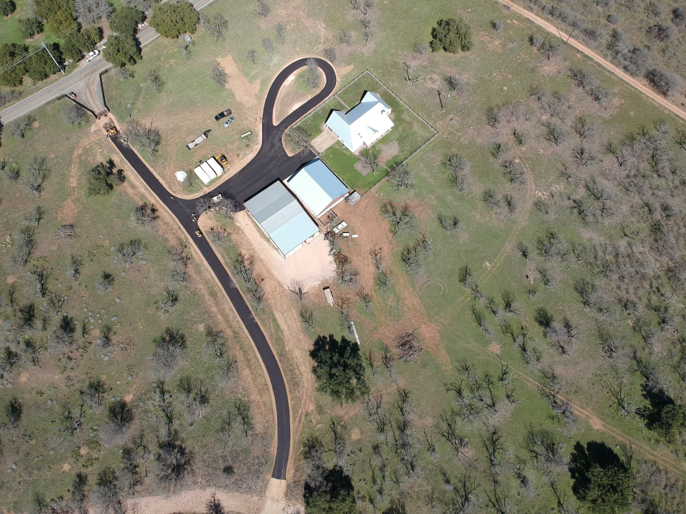 Aerial view of a rural area with a house, two metal buildings, and a paved driveway by Contractor's Asphalt, surrounded by scattered trees and grassland.