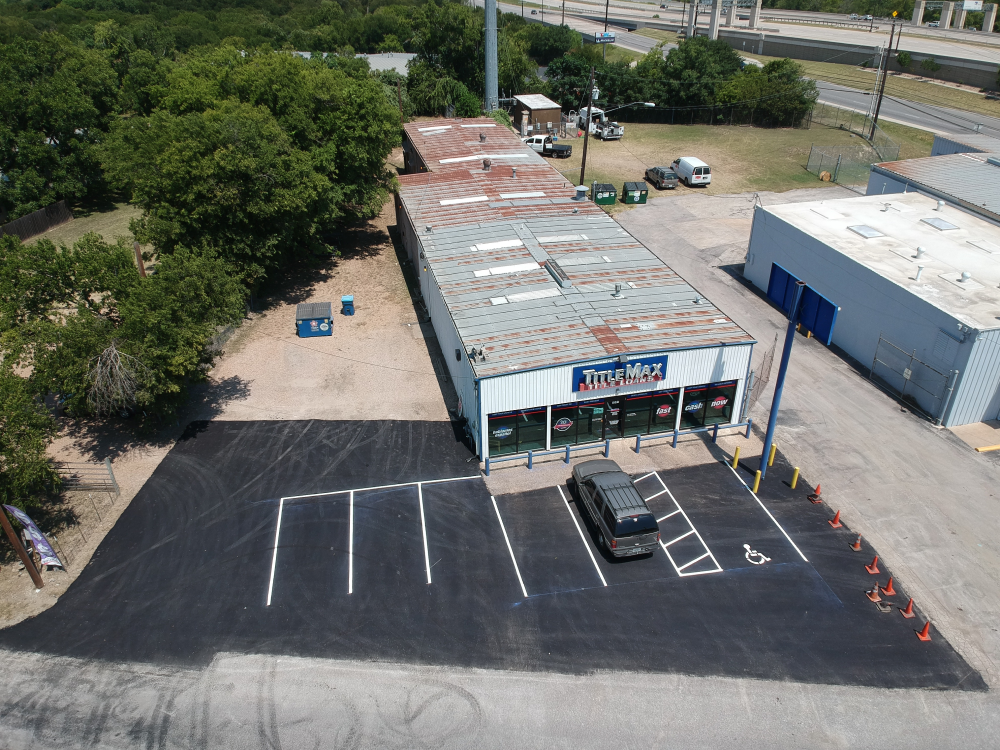 Aerial view of a business with a large parking lot, paved by Contractor's Asphalt. A single dark SUV is parked near the entrance on freshly laid asphalt. Nearby, trees line the edge of the property.