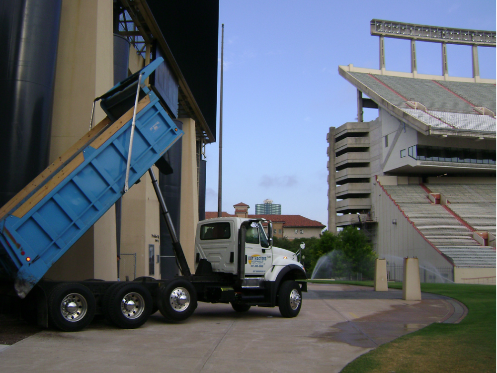 A blue dump truck from Contractor's Asphalt is unloading material next to a stadium, enhancing the paved path under a clear sky.